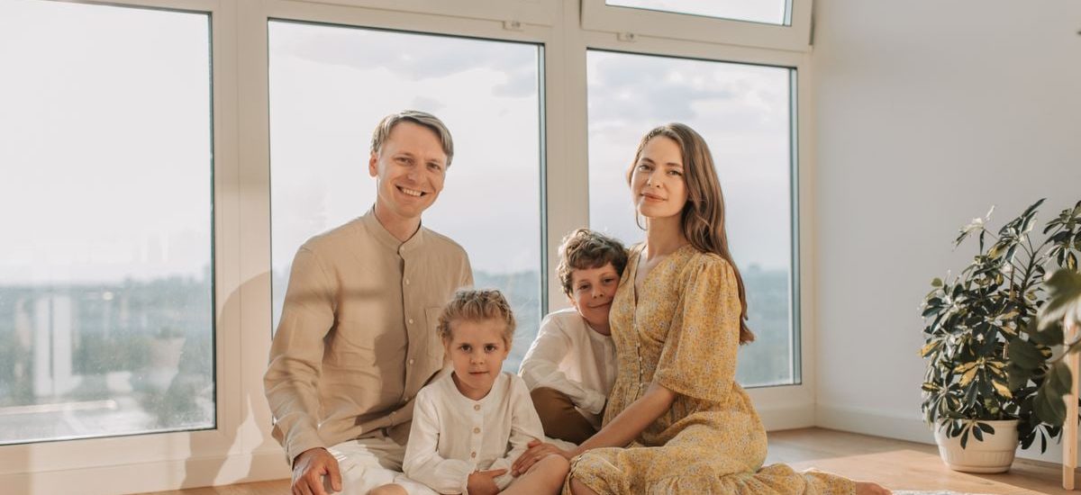 A cheerful family of four sitting together on the floor of a sunlit living room.